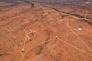 White dish shaped radio telescopes nad a large white building from the air on a red Australian desert backdrop.