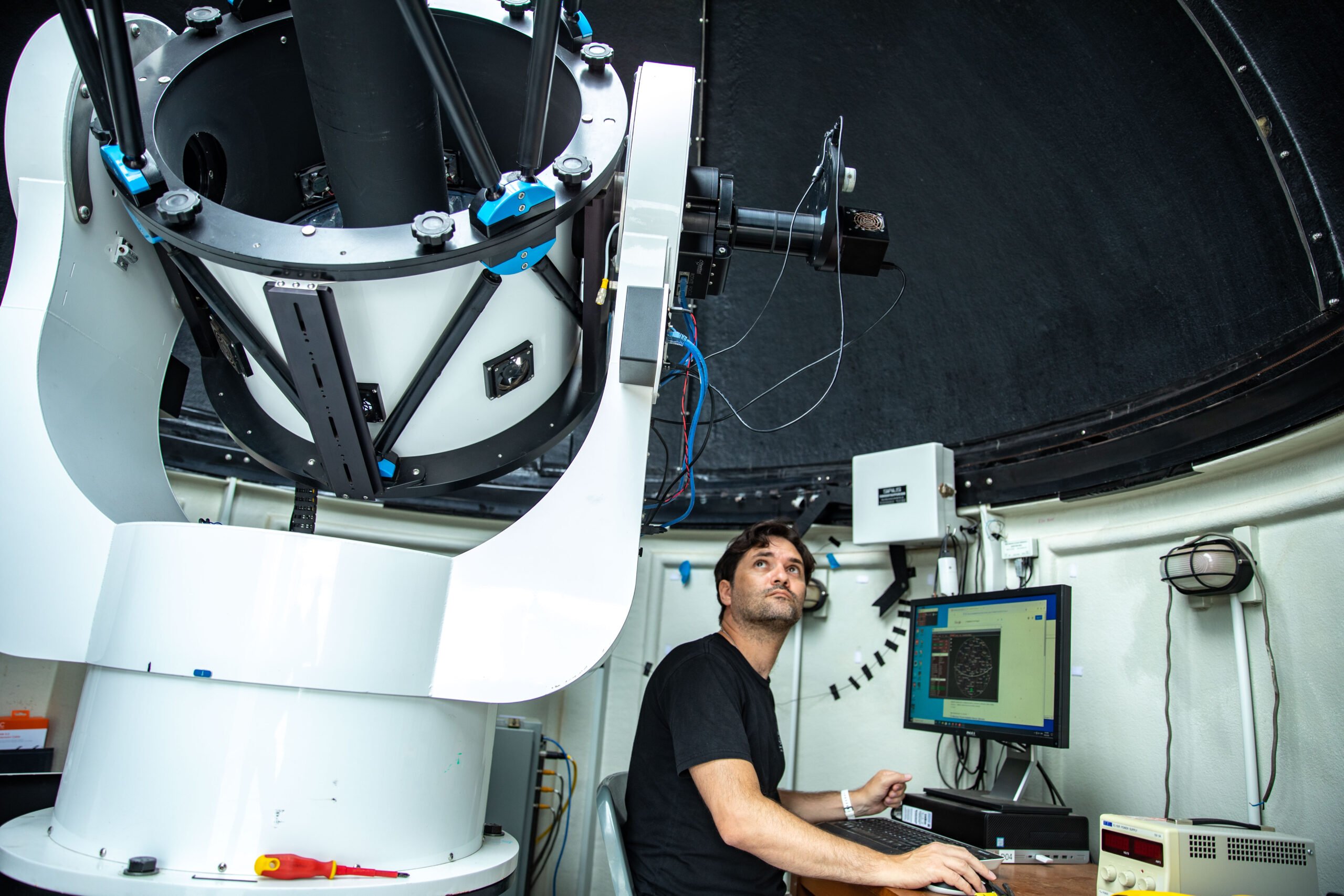 A man at a computer looks up at a large, cylindrical device on a rotating mount, pointed at the sky.