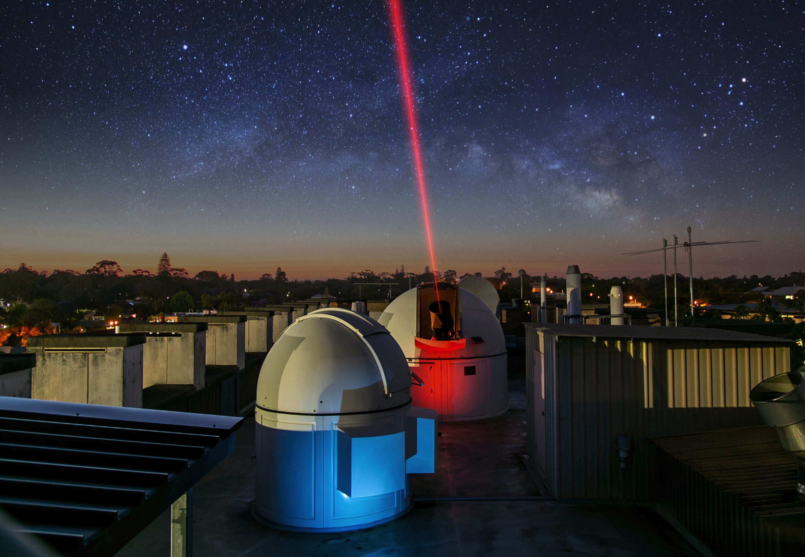 A red laser beam shines towards the sky out of an observatory dome on top of a building, with the Milky Way in the background