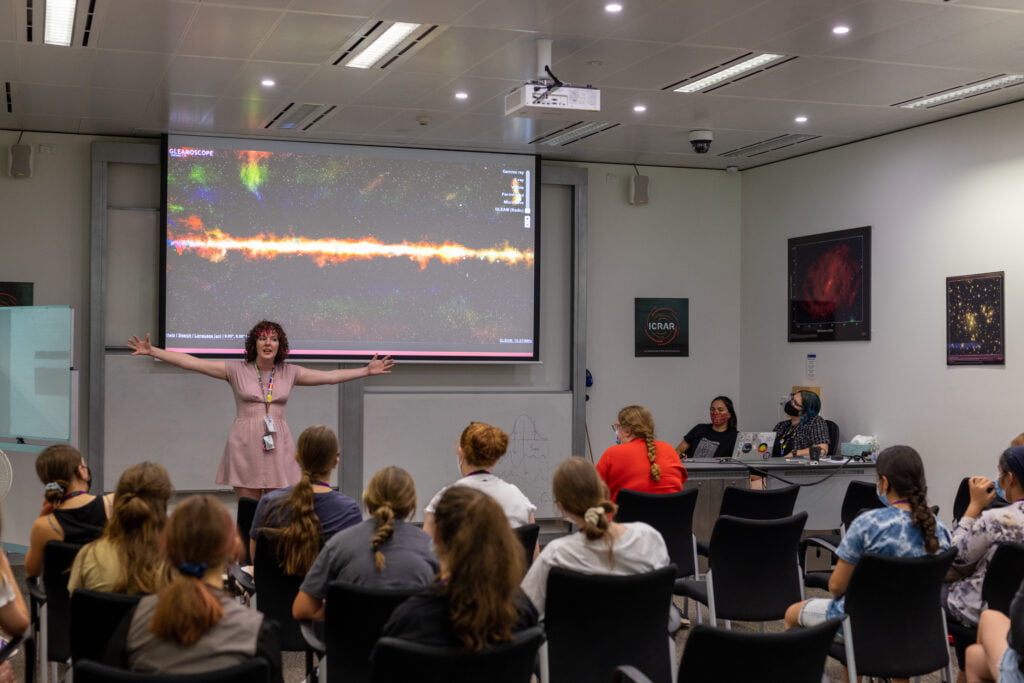 Dr Ross stands in front of an audience of female students, presenting a colourful image of a galaxy with her hands wide in excitement.