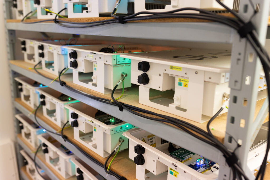 Row of metal boxes on a shelf surrounded by cables and flashing lights. 