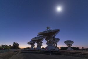 The dishes of CSIRO's ATCA telescope array against a blue sky with a bright moon in the background.
