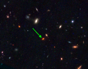 A field of several galaxies, with a particularly small red one near the centre indicated with an arrow.