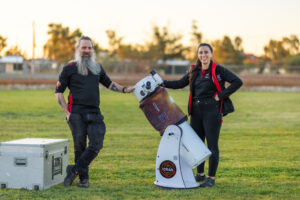 Two ICRAR team members pose with a telescope on a community oval at sunset