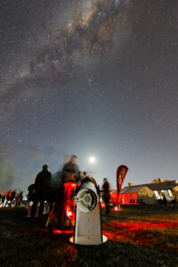 A telescope under the Milky Way, with blurred people visible in the background.